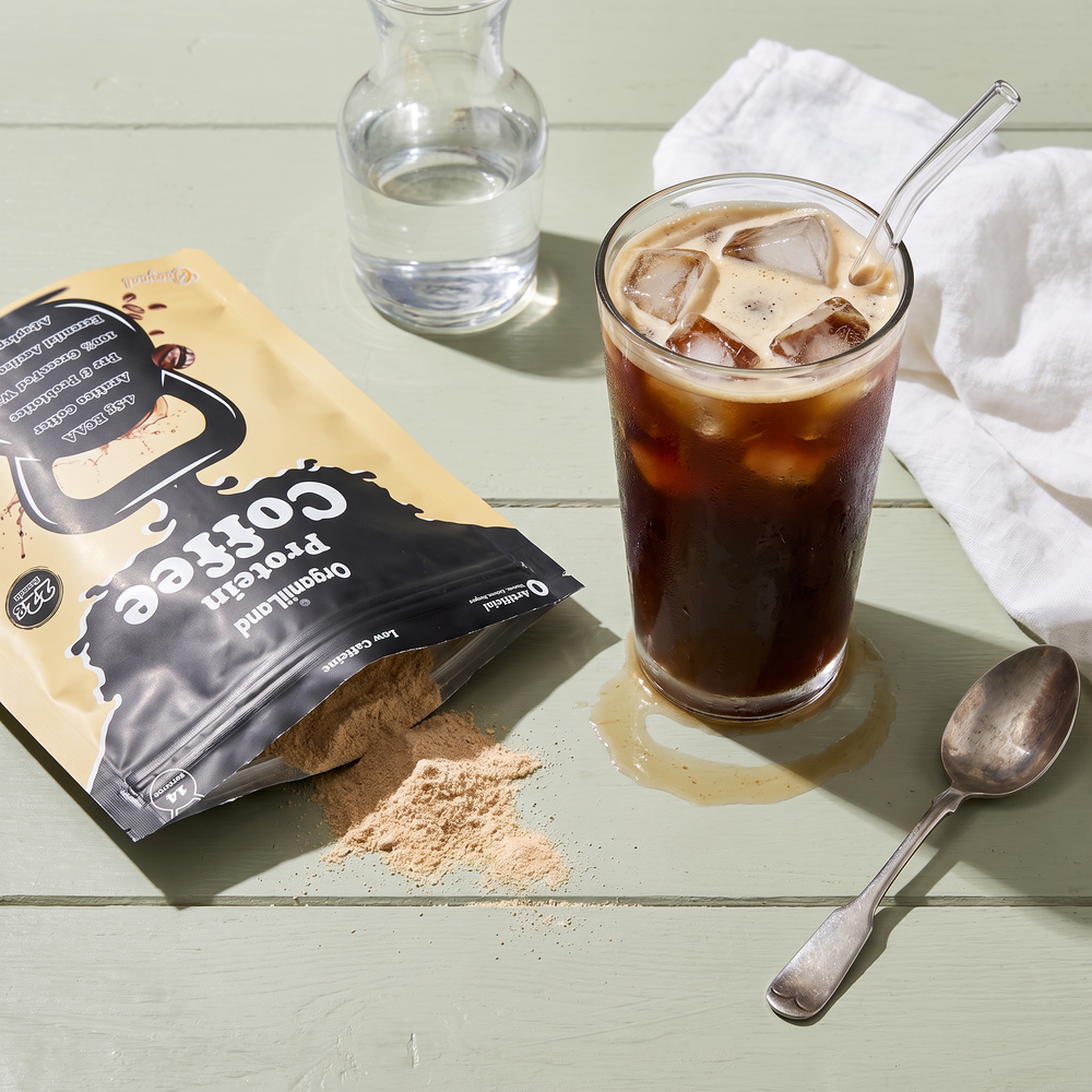 Black iced protein americano over clear ice on a sage-painted table beside an opened OrganiiLand Original Protein Coffee bag