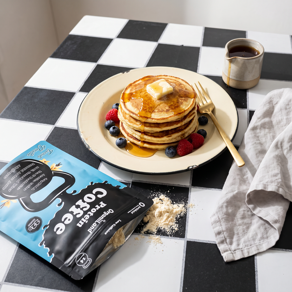 Stack of protein coffee pancakes with berries and maple on checkerboard tile beside an opened OrganiiLand French Vanilla Protein Coffee bag