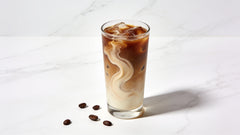 A single tall glass of iced coffee with a light cream swirl on a clean white marble surface
