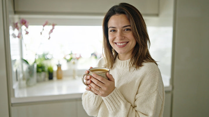 A woman in a bright kitchen holding a warm mug of coffee with a genuine smile